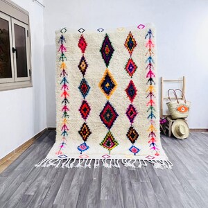 May include: Cream-colored, rectangular rug with a geometric diamond pattern in vibrant colors. The rug features a fringe along the bottom edge and colorful arrow-like designs on the sides. The diamonds are in shades of red, blue, yellow, and green.