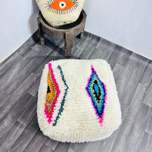May include: A white, square pouf with colourful diamond-shaped designs. A straw tote bag with an orange eye design sits on a small, carved wooden table. The bag has brown leather handles.