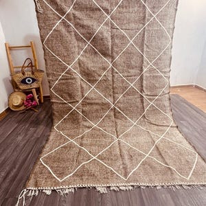 May include: A large, brown rug with a white diamond pattern. The rug has a fringed edge and is displayed against a wooden floor. A small wooden ladder and a straw bag are visible in the background.
