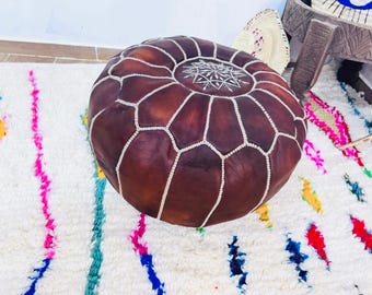 Può includere: Un pouf rotondo in pelle marrone con cuciture bianche e un design geometrico. Il pouf è posto su un tappeto bianco con motivi astratti e colorati. È un elemento d'arredo decorativo e funzionale.