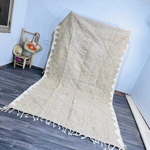 May include: A large, rectangular beige rug with a geometric diamond pattern and white fringe. The rug has a textured appearance and is placed on a dark wood floor. The rug's design includes a zigzag border.