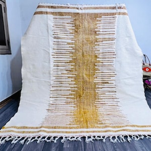 May include: A white and gold woven rug with a central vertical design and horizontal stripes. The rug has a fringed edge and is displayed on a dark wood floor. The design features a mix of white and gold tones.