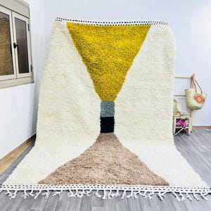 May include: A large, geometric rug with a cream-colored background and a central design in yellow, brown, black, and green. The rug has a shaggy texture and a fringe along the bottom edge. The rug is displayed on a gray wooden floor.