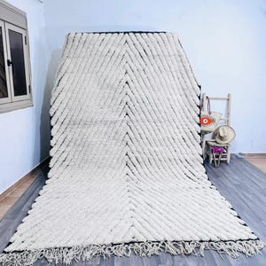 May include: A large, white rug with a black border and a chevron pattern. The rug has a shaggy texture and a fringe along the bottom edge. The rug is displayed on a gray wooden floor.