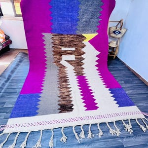 May include: A vibrant, rectangular rug with a bold geometric design. The rug features a striking combination of colors, including fuchsia, blue, white, gray, and brown. It has a fringed edge and is likely made of woven fabric.