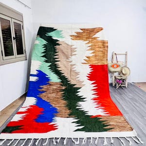 May include: A large, colorful rug with a geometric design. The rug features a white background with sections of red, blue, green, brown, and tan. The rug has a fringe along the bottom edge.
