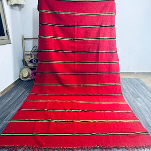 May include: A vibrant red rug with horizontal stripes in black and green. The rug has a fringed edge and is displayed against a neutral background. The rug is a decorative textile.