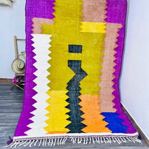 May include: A vibrant, rectangular rug with a geometric design. The rug features a bold color palette including purple, yellow, white, brown, orange, and blue. The edges have a zig-zag pattern and a fringe along the bottom. The rug is likely made of woven fabric.