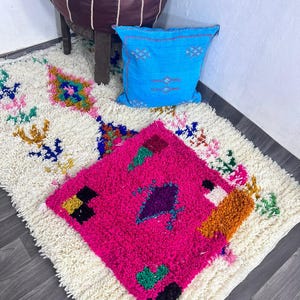 Könnte beinhalten: Ein marokkanischer Teppich mit cremefarbenem Hintergrund und bunten geometrischen Mustern. Ein leuchtend pinkfarbener quadratischer Teppich mit Rautenmuster liegt obenauf. Ein blaues Kissen und ein brauner Lederpouf sind ebenfalls im Bild.