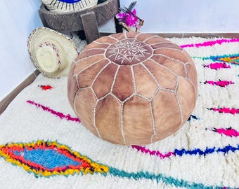 Può includere: Un pouf rotondo in pelle marrone con un motivo geometrico cucito con filo bianco. Il pouf è su un tappeto bianco con disegni astratti colorati. Sono visibili anche un cappello di paglia e un cesto intrecciato.