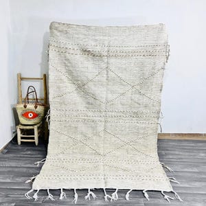 May include: A large, off-white rug with a geometric diamond pattern and horizontal lines. The rug has a fringe along the bottom edge. A small woven basket with an eye design and a wooden chair are in the background.