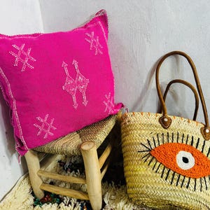 May include: A hot pink decorative pillow with white embroidered designs rests on a small wooden stool. A woven straw tote bag with brown leather handles and an orange eye design sits nearby. The items are on a patterned rug.