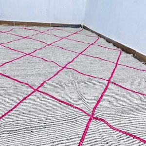 May include: A rectangular rug with a gray and white striped pattern and bright pink diamond grid design. The rug is made of a woven material and is placed on a light-colored floor.