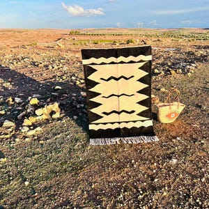 May include: A black and cream-colored geometric patterned rug with fringe is displayed outdoors. A woven straw tote bag with an eye design sits nearby. The rug is rectangular and features a zigzag design. The background shows a field under a blue sky.