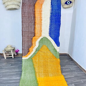 May include: A colorful, abstract rug with a geometric design. The rug features vertical stripes of brown, orange, white, and blue, with a yellow and green geometric shape at the bottom. The rug is made of a textured material and has a fringe along the bottom edge.