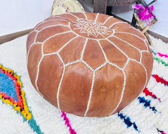 Può includere: Un pouf rotondo in pelle marrone con cuciture bianche e un motivo geometrico sulla parte superiore. Il pouf è appoggiato su un tappeto bianco a strisce colorate. Oggetti decorativi sono sullo sfondo.