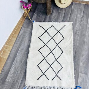 May include: A white rug with a black diamond pattern and fringe. A straw tote bag with an eye design sits on a wooden table, along with a straw hat. The rug is a decorative item.