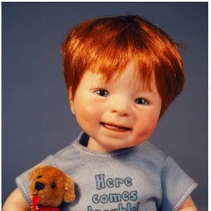 Puede incluir: Un muñeco de niño con el pelo rojo que lleva una camiseta azul con el texto "Here comes trouble!" y pantalones cortos de cuadros. Está sosteniendo un cachorro de peluche marrón con un collar rojo.