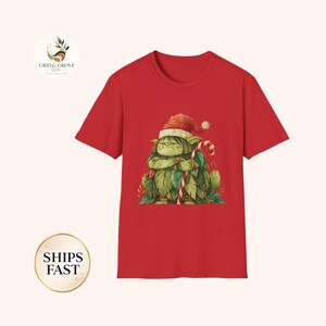 May include: A bright red short-sleeved t-shirt featuring a graphic print of a grumpy green creature wearing a red and white Santa hat. The creature is covered in green leaves and is holding a red and white striped candy cane in its arms.