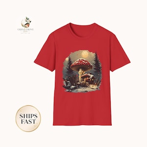 Op de afbeelding: Een felrood T-shirt met korte mouwen en een centrale grafische print van een grote rood-witte paddenstoel in een besneeuwd boslandschap, met een klein huisje eronder, omringd door donkere dennenbomen en een gloeiende volle maan.