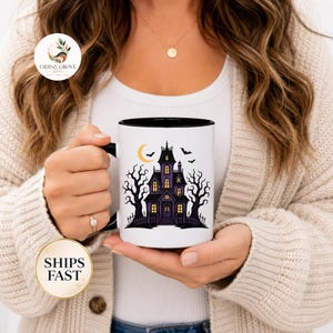 Puede incluir: Una taza de cerámica blanca con borde y asa negros, que presenta una ilustración gráfica de una casa encantada morada, árboles negros sin hojas, una luna creciente amarilla y murciélagos volando sobre un fondo blanco.