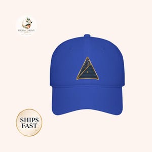 Puede incluir: Una gorra de béisbol azul real con un emblema triangular centrado. El emblema tiene un borde dorado y contiene un campo azul oscuro con un patrón de pequeñas estrellas blancas en la parte superior y rayas horizontales en la inferior.