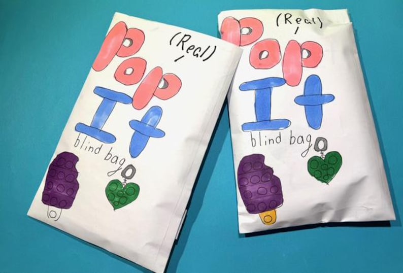Pu&ograve; includere: Due sacchetti ciechi bianchi "Pop It" con testo e illustrazioni disegnate a mano. Le borse presentano scritte rosa e blu, un ghiacciolo viola e un ciondolo a forma di cuore verde. Le parole "(Real)" sono scritte sopra.