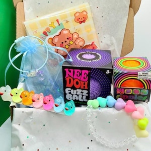 May include: A gift box containing a Nee Doh Fuzz Ball box, a Nee Doh Ripples box, a light blue organza bag with a character bracelet, a row of small colorful plastic ducks, a pastel heart-shaped bead bracelet, and a card with a cartoon bear.