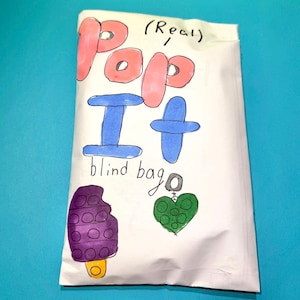 Pu&ograve; includere: Un sacchetto di carta bianco con le parole "POP IT blind bag" in lettere colorate disegnate a mano. La borsa presenta illustrazioni di un ghiacciolo viola, un cuore verde e un disegno blu a forma di croce. La parola "(Real)" &egrave; scritta sopra la parola "POP".