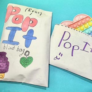 Pu&ograve; includere: Due sacchetti di carta bianchi con le parole "Pop It" scritte sopra. Un sacchetto &egrave; etichettato "blind bag" e presenta disegni di un ghiacciolo, un cuore e un ciondolo. L'altro sacchetto ha un giocattolo Pop It a forma di cuore che sbuca.