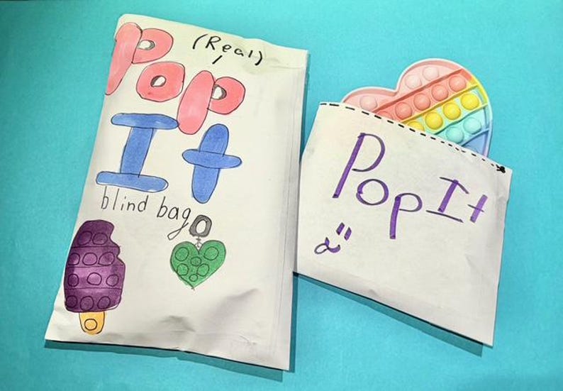 Pu&ograve; includere: Due sacchetti di carta bianchi con le parole "Pop It" scritte sopra. Un sacchetto &egrave; etichettato "blind bag" e presenta disegni di un ghiacciolo, un cuore e un ciondolo. L'altro sacchetto ha un giocattolo Pop It a forma di cuore che sbuca.