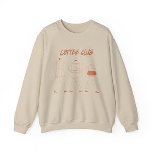 Puede incluir: Una sudadera de cuello redondo en color beige con un estampado gráfico en tono marrón rojizo. El diseño muestra el texto COFFEE CLUB sobre tres cafeteras estilizadas, cada una representada con piernas y pies finos, caminando en una fila.