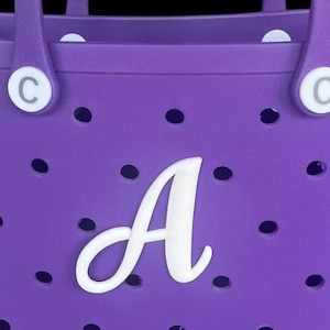 Könnte beinhalten: Nahaufnahme einer lila Tragetasche mit einem weißen Buchstaben "A" auf der Vorderseite. Die Tasche hat ein perforiertes Design und zwei Griffe. Der Buchstabe "C" ist oben auf der Tasche sichtbar.