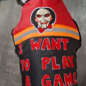 May include: A dog wearing a black and red vest with the text "I WANT TO PLAY A GAME" in red letters. The vest has a graphic of a scary face and the Kong logo.