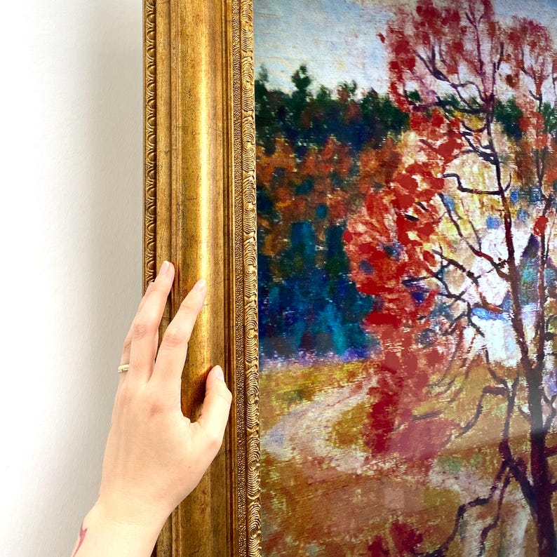 May include: A framed landscape painting with a gold ornate frame. The artwork depicts an autumn scene with a red tree, a path, and a forest in the background. The frame is held by a hand.