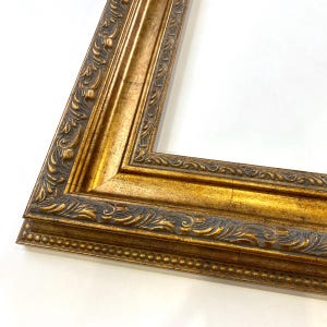 May include: Ornate gold picture frame with detailed carvings and a dark gray accent. The frame has a classic, vintage aesthetic, with a layered design and a beaded edge. The corner of the frame is visible against a white background.