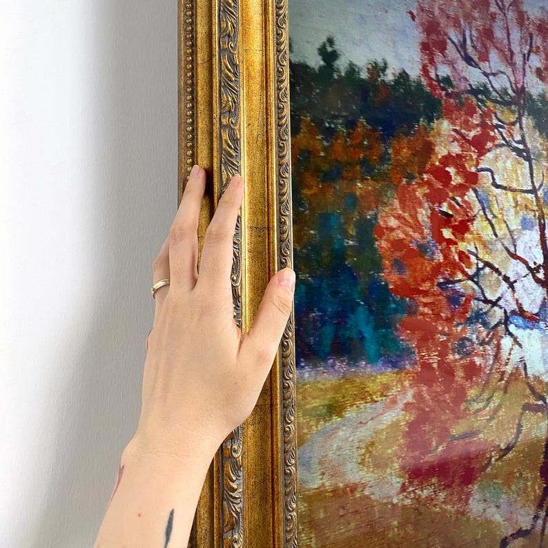 May include: A hand touches a gold-colored, ornate picture frame. The framed artwork features a landscape painting with a path, trees, and a vibrant display of red and orange foliage. The painting's colors contrast with the gold frame.
