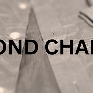 May include: Close-up of a light gray button-down shirt with the text "SECOND CHAPTER" in black. The shirt is partially unbuttoned, with the cuff of the sleeve folded over, showing a button.