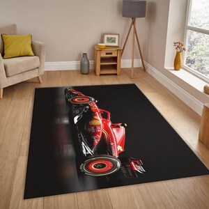 May include: A rectangular black area rug featuring a red Formula 1 race car design. The car has black and red accents, with visible sponsor logos. The rug is placed on a hardwood floor, with a neutral-toned living room in the background.