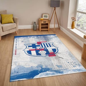 May include: A rectangular area rug featuring the FC Barcelona crest in blue, red, and white. The rug has a distressed, artistic design with blue paint splatters and the text "MES QUE UN CLUB FC BARCELONA" along the side. The rug is placed on a wooden floor.