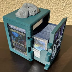 May include: A teal 3D-printed deck box with a gray helmet-shaped emblem on top. The box features a side-loading compartment holding a stack of trading cards and a front-facing display slot containing a single card protected by a clear plastic case.