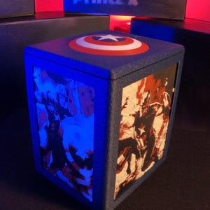 May include: A blue cubic lamp featuring a Captain America shield graphic on the top surface. The side panels display comic book illustrations of superheroes in red, white, and blue tones. The structure has a textured finish and emits a blue light.