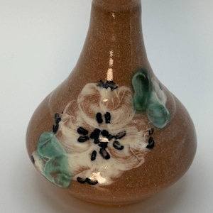 May include: A small, bulbous ceramic vase with a speckled brown glaze. The body features a hand-painted floral design in white with black accents and green leaves. The vase has a narrow neck and a flared rim, showcasing a smooth, glossy finish.