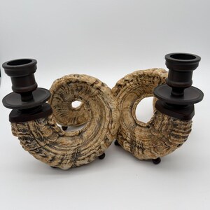 May include: A pair of candle holders featuring a textured, tan and brown spiral horn-shaped base. Each holder is topped with a dark brown, cylindrical candle cup and a circular drip plate, supported by small dark feet on a plain white background.