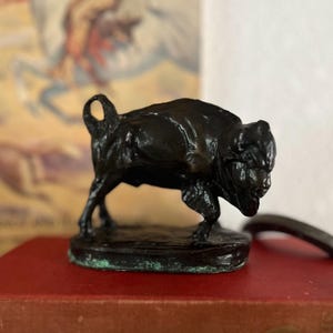 May include: A small, dark bronze sculpture of a bison, standing on a circular base. The sculpture has a detailed texture and a slightly weathered appearance. The bison is depicted in a walking pose, with its head lowered. The sculpture is placed on a red surface.