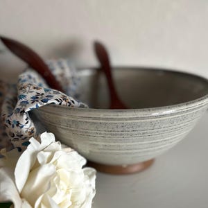 Puede incluir: Un cuenco de cerámica gris claro con base y borde marrón, con una superficie texturizada. Dos cucharas de madera descansan dentro del cuenco, con un paño con estampado floral sobre el lateral. Una rosa blanca está en primer plano.