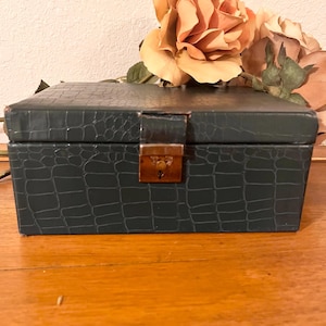 May include: A dark teal jewelry box with a faux crocodile skin texture. The rectangular box has a hinged lid and a gold-colored clasp. A cluster of peach-colored roses sits on top of the box.
