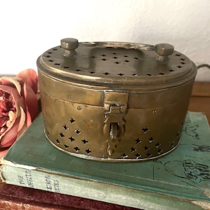 May include: A vintage brass oval box with a hinged lid and a latch closure. The box features decorative star-shaped cutouts around the sides and top. Two small knobs are on the top. The box is sitting on top of a stack of old books.