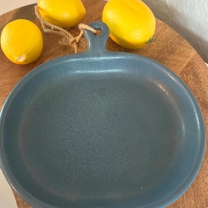 May include: A blue-green ceramic serving platter with a handle, accompanied by three yellow lemons. The platter is oval-shaped and rests on a wooden surface, with a natural rope detail. The lemons are bright yellow.