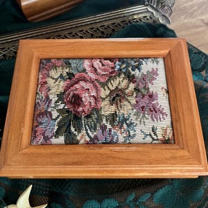 May include: A rectangular wooden jewelry box with a floral tapestry design on the lid. The tapestry features pink, red, and purple roses with green leaves. The box is made of light brown wood.